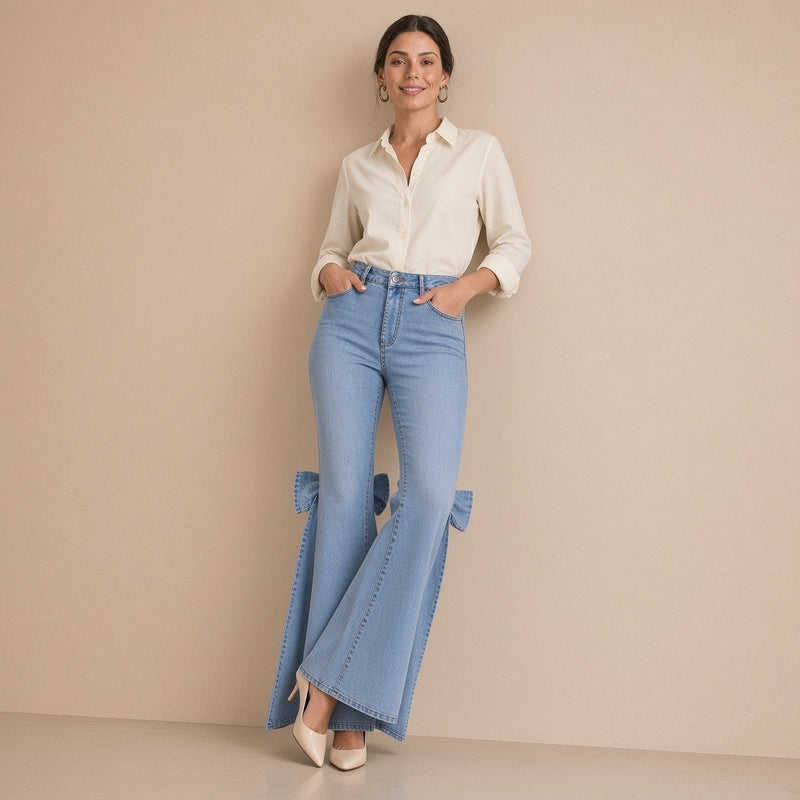 Jean flare bleu clair porté de face, appuyée contre le mur, chemise écrue rentrée : on devine les nœuds en denim derrière les genoux