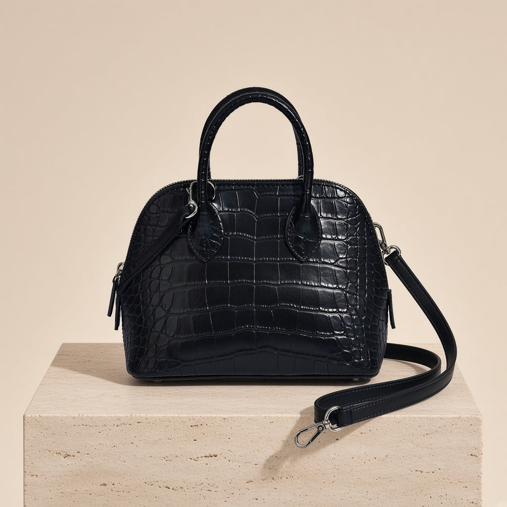 Mini sac bowling en cuir effet croco noir posé sur un socle en pierre, bandoulière amovible
