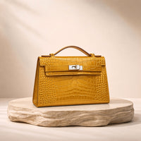 Pochette en cuir effet croco SAKIWE jaune moutarde, posée sur un socle en pierre
