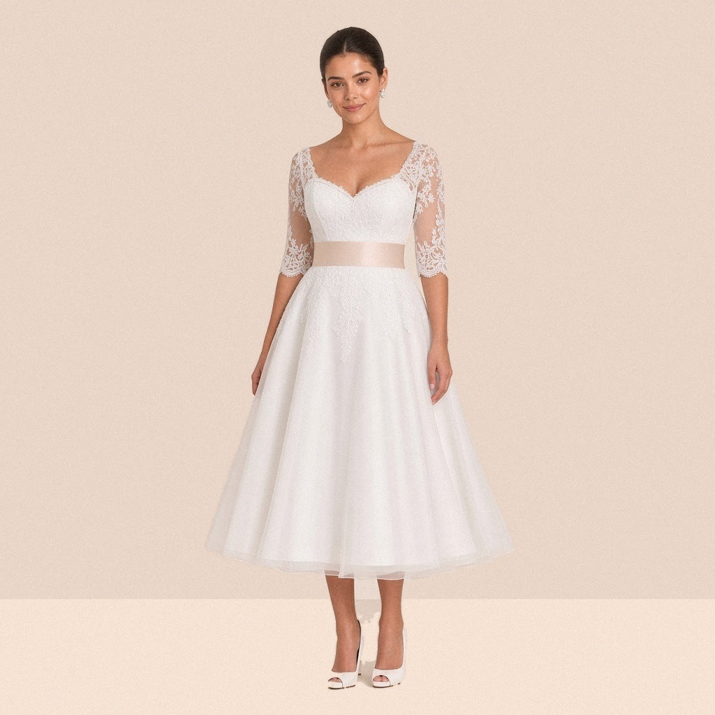 Robe de mariée blanche mi-longue en dentelle portée de face, manches au coude et ceinture satinée champagne