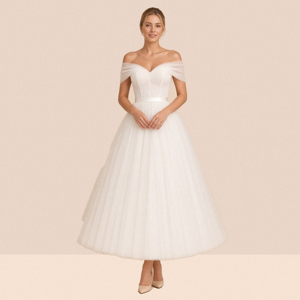 Robe de mariée ivoire mi-longue en tulle à épaules dénudées et ceinture satinée – vue de face, mains jointes à la taille.