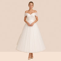 Robe de mariée ivoire mi-longue en tulle à épaules dénudées et ceinture satinée – vue de face, mains jointes à la taille.