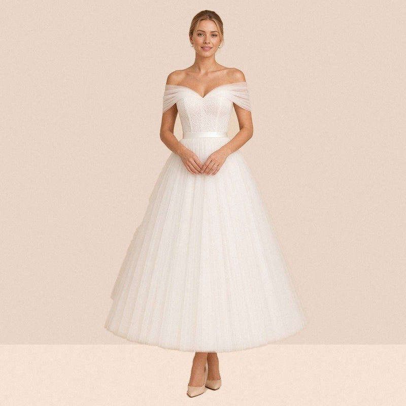 Robe de mariée ivoire mi-longue en tulle à épaules dénudées et ceinture satinée – vue de face, mains jointes à la taille.