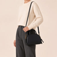 Gros plan du sac bowling demi-lune noir porté à l’épaule, détail de la poche plaquée et des mousquetons